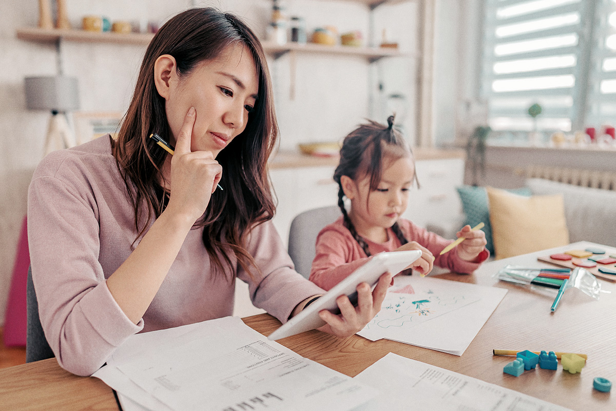 Woman looking at tablet sitting at table while child draws next  to her